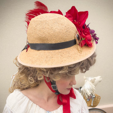 Woman wearing the Hair Helmet 1789, a bicycle helmet with a curly wig-style cover and decorated straw hat featuring red flowers and feathers